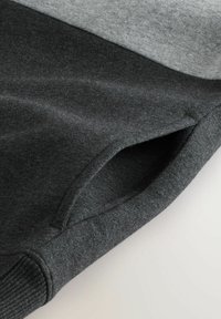 Dunkelgrauer Sweatshirt-Ärmel mit Rippenbündchen und schrägem Seitentaschendetail auf hellgrauem Stoffhintergrund.