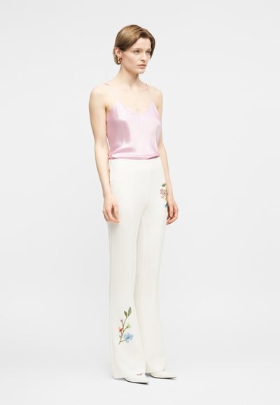 Γυναίκα που στέκεται, φοράει ένα ανοιχτό ροζ σατέν καμιζόλ και άσπρο παντελόνι με φαρδιά μπατζάκια, διακοσμημένο με πολύχρωμη κεντητή floral κοντά στο τελείωμα και στο μηρό.