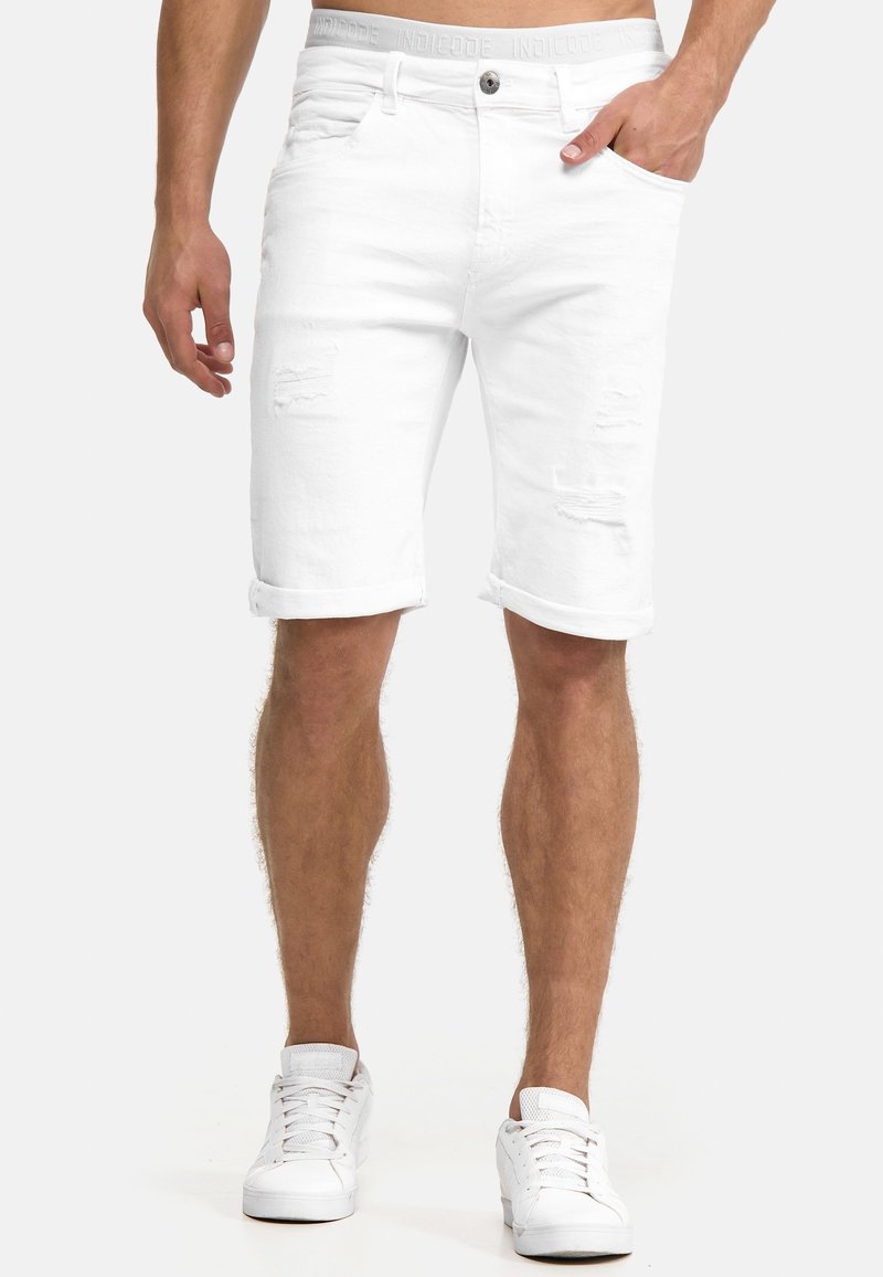 Homme portant un short en denim blanc à ourlet retroussé avec des détails usés, et des baskets blanches, debout avec une main dans la poche.