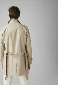 Beżowy płaszcz trench z paskiem, z tylnym rozporkiem i klasycznym kołnierzem. Tkanina wydaje się gładka, odpowiednia do warstwowych stylizacji.