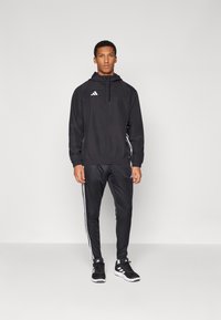 Giacca sportiva nera con cappuccio e zip, con un logo bianco. Abbinata a pantaloni neri e scarpe da ginnastica nere con dettagli bianchi.