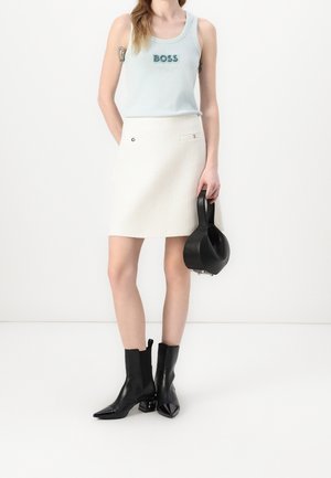 Femme portant un débardeur bleu clair avec le texte "BOSS", une jupe blanche, des bottines noires et tenant un sac à main noir sur un fond blanc.