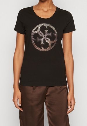 Personne portant un t-shirt noir cintré avec un motif rond, scintillant et entrelacé sur la poitrine et un pantalon en satin marron.