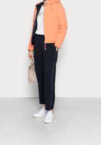 Persona che indossa una giacca con cappuccio color corallo, una t-shirt bianca con grafica, pantaloni neri con coulisse, scarpe da ginnastica bianche e tiene in mano una borsa beige.