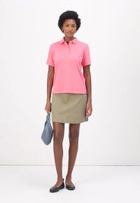 Roze polo-shirt met korte mouwen, gecombineerd met een olijfgroene rok tot de knie en zwarte ballerina's. Het model houdt een lichtblauwe handtas vast.