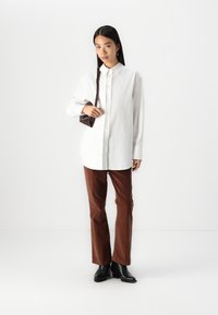 White button-up shirt, brown corduroy trousers, and black ankle boots. Model holds a small brown handbag. Simple, structured design.