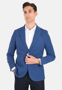 Blazer blu texturizzato con rever a scalinato, chiusura a un bottone e due tasche frontali. Camicia bianca sotto, con fazzoletto da taschino.