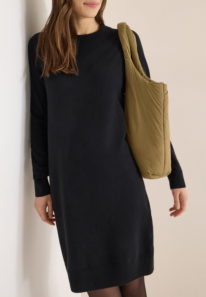 Robe en maille noire à manches longues, avec poignets et ourlet côtelés. Présentant un sac d'épaule matelassé vert. Texture lisse, coupe longueur genou.