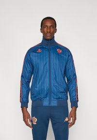 Ναυτικό μπλε τζάκετ φόρμας με κάθετες ρίγες, ψηλό γιακά και κόκκινες λεπτομέρειες· διαθέτει λογότυπο Kappa και έμβλημα ομάδας στο στήθος.