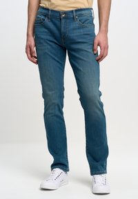 Jeans de mezclilla azul de corte slim con secciones desgastadas, cinco bolsillos, cierre de botón y costuras en contraste. Combinados con zapatillas blancas.