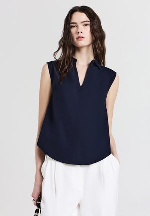 Donna con lunghi capelli mossi che indossa una blusa senza maniche di colore blu navy e pantaloni bianchi, mentre tiene una borsa di perline bianca e nera contro uno sfondo semplice.