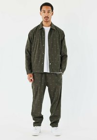 Completo giacca e pantaloni verde scuro abbinati con un motivo logo sottile, realizzato in un materiale liscio, indossato con una t-shirt bianca e sneakers.