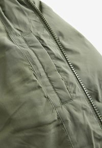 Jachetă căptușită de culoare verde măslin, cu un fermoar diagonal și o buzunar frontal, având o textură netedă și detalii de cusătură curate.