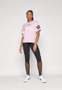Lyserød kortærmet t-shirt med sort Adidas-logo og tre striber på ærmerne, parret med sorte leggings med mesh-detaljer og hvide sko.