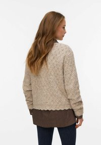 Suéter beige de punto con un patrón de diamantes texturizado y un dobladillo festoneado, llevado sobre una camisa marrón a rayas y vaqueros oscuros.