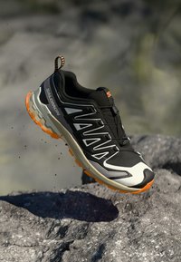 Black and orange Salomon trail running shoe mid-air above rocky terrain with dirt particles visible around the sole.