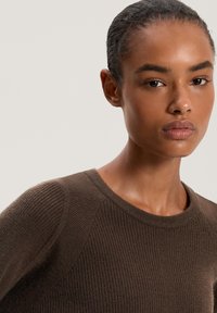 Maglione marrone a coste con collo rotondo e maniche raglan, caratterizzato da una consistenza morbida e una silhouette aderente. Primo piano del viso di una modella.