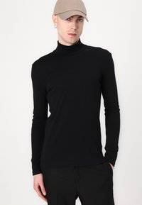 Camisa negra de manga larga y cuello vuelto, fabricada con un tejido suave, presenta un diseño ajustado y una textura lisa, combinada con pantalones negros.