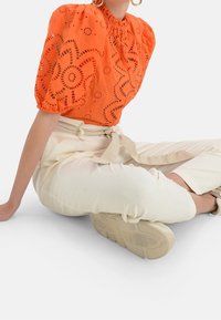 Oranje blouse met opgediepte mouwen, voorzien van uitgesneden bloemmotieven. Gecombineerd met crèmekleurige tapered broek en beige sneakers.