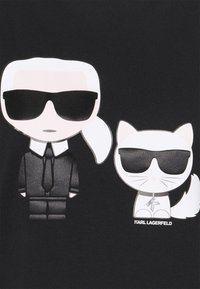 Černá látka má kreslený motiv postavy v obleku a slunečních brýlích vedle bílé kočky se slunečními brýlemi. Text zní "KARL LAGERFELD."