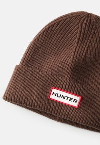 Hnedá rebrovaná pletená čiapka s prehnutým okrajom, so štvorcovou nálepkou s červeným okrajom a čiernym textom "HUNTER."