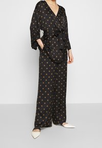 Black satin pajama set with a wrap-style top and wide-leg pants, featuring a gold circular pattern and tied waist detail. Pointed white shoes.
