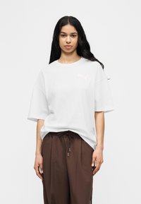 Νεαρός/ή με μακριά μαύρα μαλλιά φοράει φαρδιά λευκή μπλούζα και καφέ παντελόνι με κορδόνι, στέκεται μπροστά σε απλό ανοιχτό φόντο.