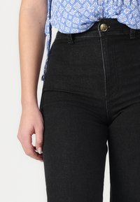 Jean noir taille haute au design ajusté, fabriqué en denim. Comprend un bouton en laiton et une couture contrastante discrète.