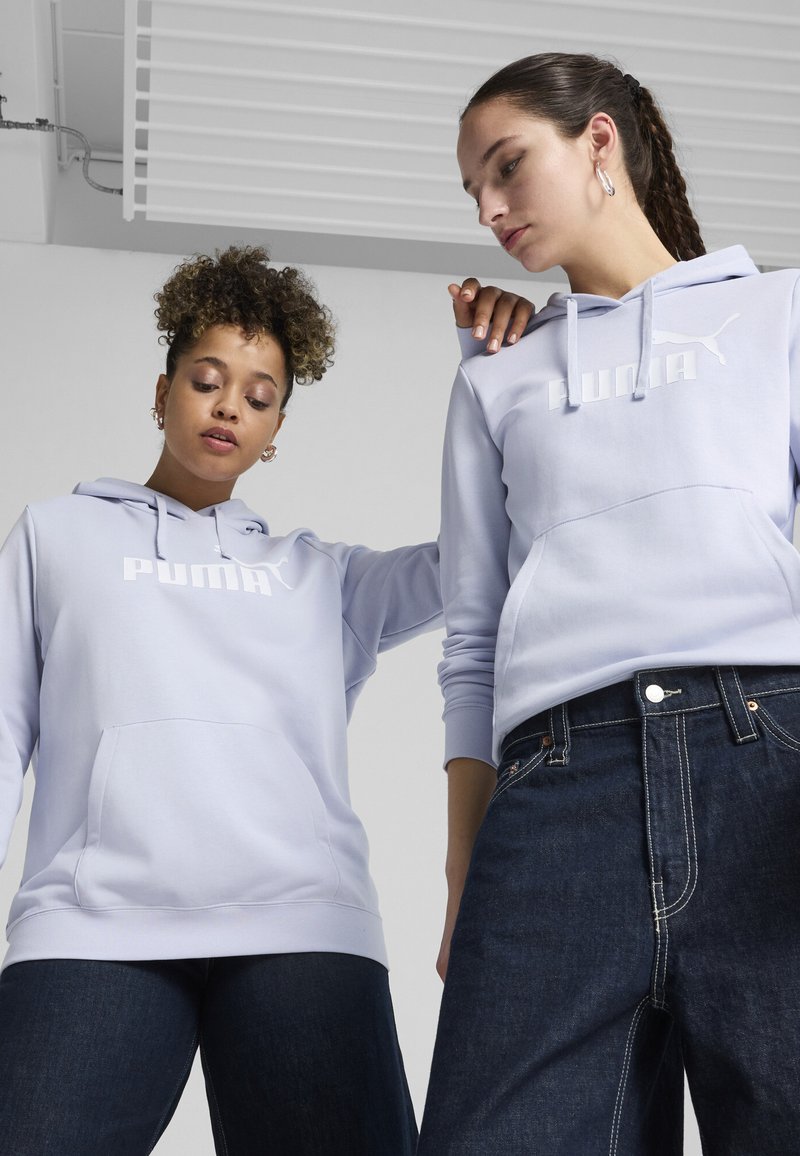 Jasnofioletowe bluzy z kapturem z białym logo Pumy i przednimi kieszeniami, zestawione z ciemnoniebieskimi dżinsami. Gładki materiał i luźny krój.