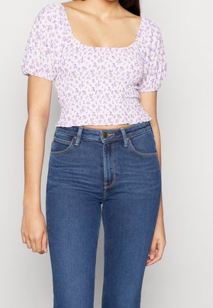 Modèle portant un haut blanc à imprimé floral violet, avec des manches bouffantes et une encolure carrée, associé à un jean bleu taille haute.
