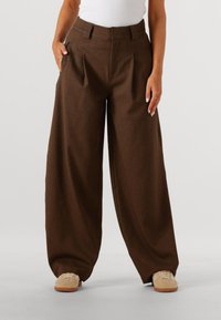 Bruine wijde broek met plooien, voorzien van zijzakken en een gladde textuur. Gecombineerd met beige sneakers.