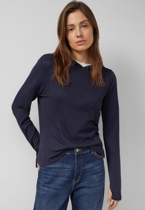 Femme aux cheveux bruns raides, portant un haut à manches longues bleu marine et un jean bleu, debout avec une main derrière le dos sur un fond uni.