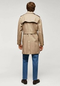 Beige trenchcoat met een ceintuur in de taille, dubbelzijdig ontwerp, schouderepauletten en een achterventilatie, gecombineerd met blauwe denim jeans en donkere schoenen.