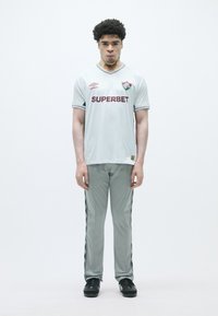 Camisa de futebol cinza claro com acentos em marrom e logótipo; combinada com calças cinzentas correspondentes apresentando riscas laterais e ténis pretos.