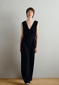 Svart uten ermer jumpsuit med dyp V-hals og bred benutforming. Laget av glatt stoff med en subtil glans, med minimalt med tilbehør.