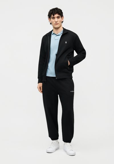 Muž s kudrnatými tmavými vlasy, oblečený v světle modrém polokošili, černé bundě na zip, černých teplácích a bílých teniskách, stojící s jednou rukou v kapse.