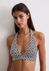 Mujer con un top halter en blanco y negro con un patrón cuadrado de flores, de pie contra una pared blanca lisa.
