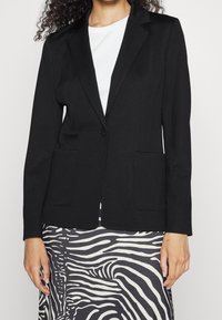 Blazer nero in tessuto liscio, con un solo bottone, rever a punta e due tasche frontali, indossato sopra una gonna dal motivo fantasia.