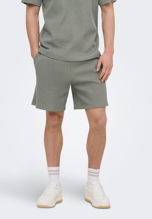 Mand iført matchende grå teksturerede shorts og kortærmet top, hvide crew-sokker og hvide sneakers, stående med hænderne i lommerne.