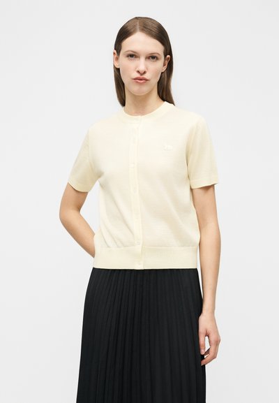 Γυναίκα με ίσια καστανή τρίχα φοράει ένα κοντομάνικο εκρού κουμπωτό ζακέτα και μια μαύρη πλισέ φούστα, στέκεται μπροστά σε απλό φόντο.