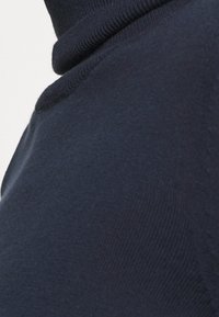 Läheltä kuvattu tummansininen neulottu korkeakauluksinen villapaita, jossa on ribbauskaulus ja teksturoitu kangas olkapäällä ja kaulassa.