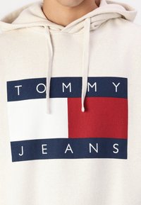Beige hoodie with a drawstring, featuring a large graphic in navy, white, and red blocks, with "TOMMY JEANS" text in bold white letters.