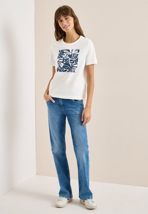Vrouw staat glimlachend met haar hand in haar zak, ze draagt een wit t-shirt met een blauw abstract vierkant ontwerp, een blauwe spijkerbroek en witte sneakers.