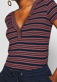 Granatowy body z prążkowanej tkaniny w koralowe i białe paski, z dekoltem w kształcie litery V oraz trzema brązowymi guzikami. Połączone z ciemnoniebieskimi spodniami na sznurki.