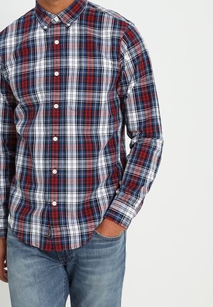 Mężczyzna z ręką w kieszeni, ubrany w czerwoną, białą i niebieską koszulę w kratę z długim rękawem zapinaną na guziki oraz jasnoniebieskie dżinsy na jednolitym tle.