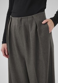 Person, der weite graue plissierte Hosen und ein schwarzes Langarmoberteil trägt, mit der rechten Hand in der Hosentasche.