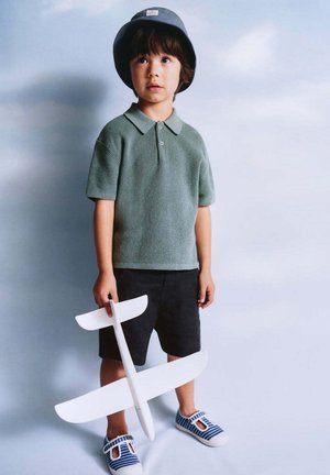 Jeune garçon avec un chapeau bob et un polo vert tenant un avion jouet blanc, debout sur un fond bleu clair.