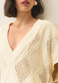 Kremast pleten pulover z globokim V-izrezom, ki vsebuje teksturirane vzorce odprtega tkanja in rebraste poudarke ob izrezu in rokavih.