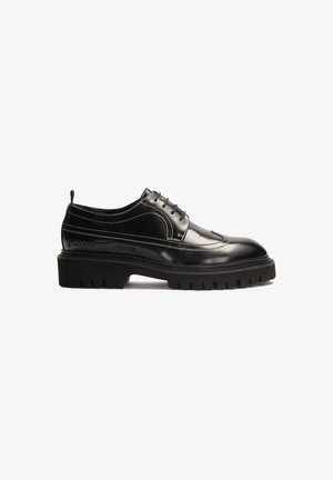 Chaussure en cuir noir avec un bout arrondi, une semelle en caoutchouc texturé épaisse, des coutures blanches contrastantes et un laçage.