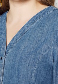 Jasnoniebieska denimowa bluzka z dekoltem w kształcie litery V, z guzikami i lekko teksturowanym materiałem. Posiada widoczne szwy wzdłuż krawędzi.
